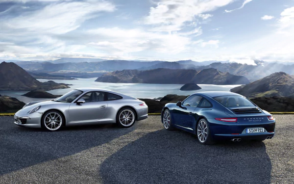 HD PC desktop wallpaper showing two Porsche 911 Carrera vehicles (silver and blue) parked on a mountain overlook above a lake under a dramatic cloudy sky.