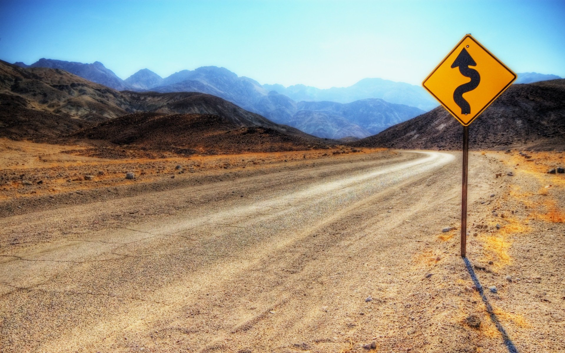 HD PC desktop wallpaper of a winding road sign on a dusty desert road with mountains in the background under a clear blue sky.