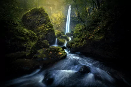 An HD desktop wallpaper featuring a serene waterfall flowing into a moss-covered river in a lush forest, bathed in soft, natural light.