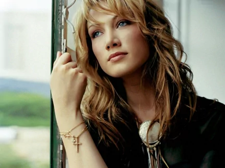 HD PC desktop wallpaper of a contemplative female singer by a window, long wavy hair, wearing a cross bracelet and pendant, soft natural lighting and muted background.