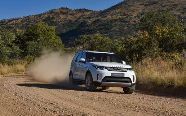 White Land Rover Discovery SUV kicking up dust while driving on a rugged dirt road with hills and greenery in the background. HD desktop wallpaper of a Land Rover vehicle.