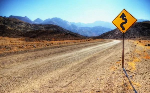 HD PC desktop wallpaper of a winding road sign on a dusty desert road with mountains in the background under a clear blue sky.
