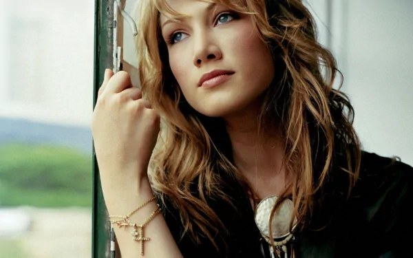 HD PC desktop wallpaper of a contemplative female singer by a window, long wavy hair, wearing a cross bracelet and pendant, soft natural lighting and muted background.