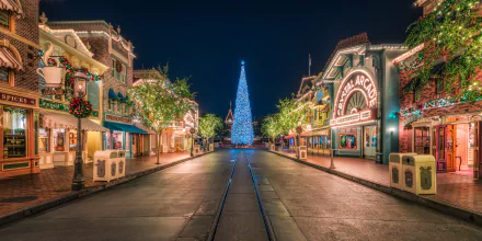 A brightly lit street at Disneyland, California, features a towering Christmas tree at its center, with festive lights adorning buildings on either side. The image serves as an HD desktop wallpaper.