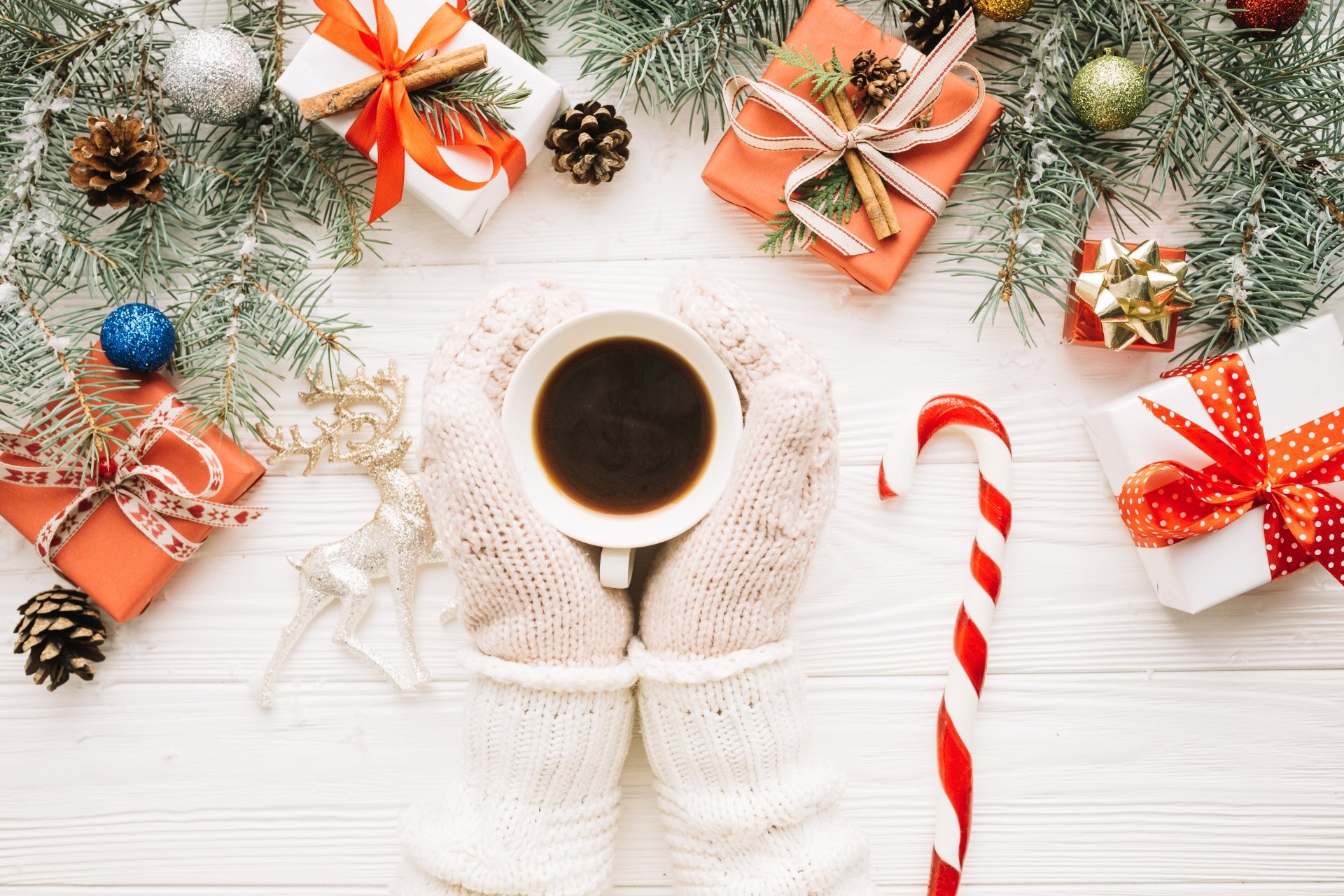 Cozy hands in white mittens holding a cup of coffee surrounded by Christmas gifts, candy cane, pine cones, and festive greenery on a white wooden background.