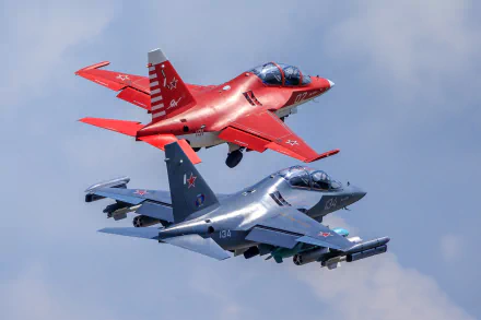 Two Yakovlev Yak-130 jet fighter warplanes, one red and one gray, fly closely against a blue sky in this HD military aircraft wallpaper.