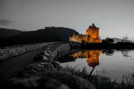 Eilean Donan Castle in Scotland illuminated at dusk, reflected clearly in the calm water, with a stone bridge leading to the historic man-made structure.