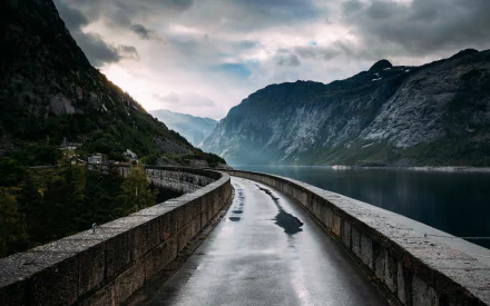 A serene fjord in Norway features a man-made dam, with a winding pathway along the water's edge surrounded by towering mountains and a moody sky.