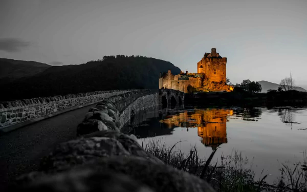 Eilean Donan Castle in Scotland illuminated at dusk, reflected clearly in the calm water, with a stone bridge leading to the historic man-made structure.