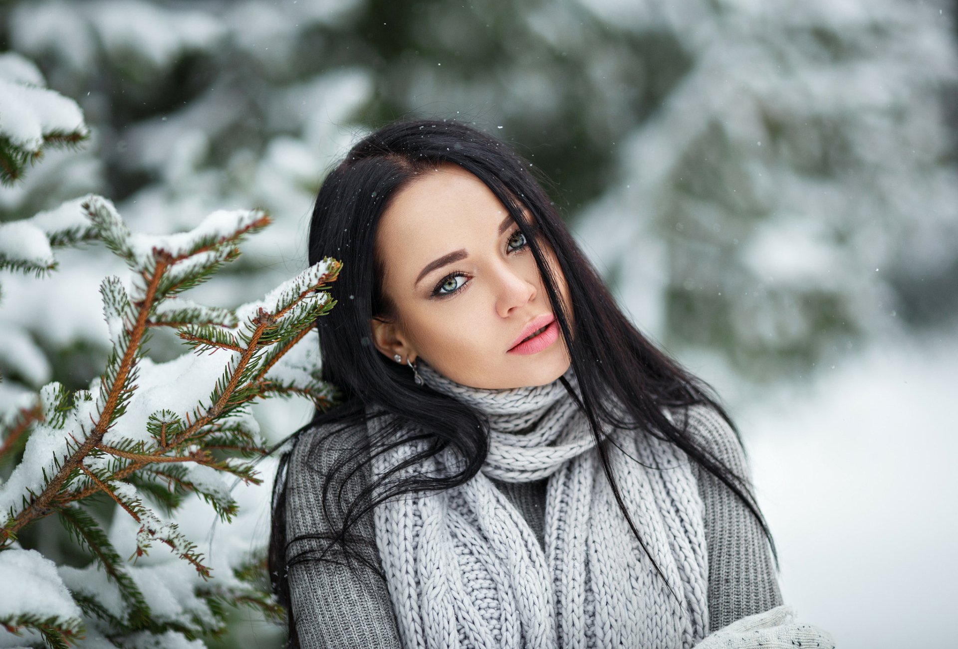 HD wallpaper of a woman with black hair and green eyes, wearing a gray scarf, standing by snow-covered pine branches in a winter forest, featuring Angelina Petrova.