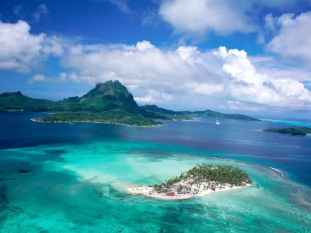 A stunning aerial view of a tropical island surrounded by crystal-clear waters, with lush greenery and soft white sand, under a bright blue sky dotted with fluffy clouds.