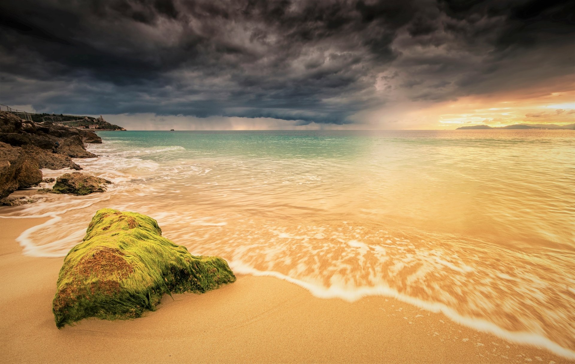HD wallpaper of a Sardinian beach in Italy, showcasing the horizon with dramatic clouds, a serene sea merging with the sandy coast, and green moss-covered rocks in the foreground.
