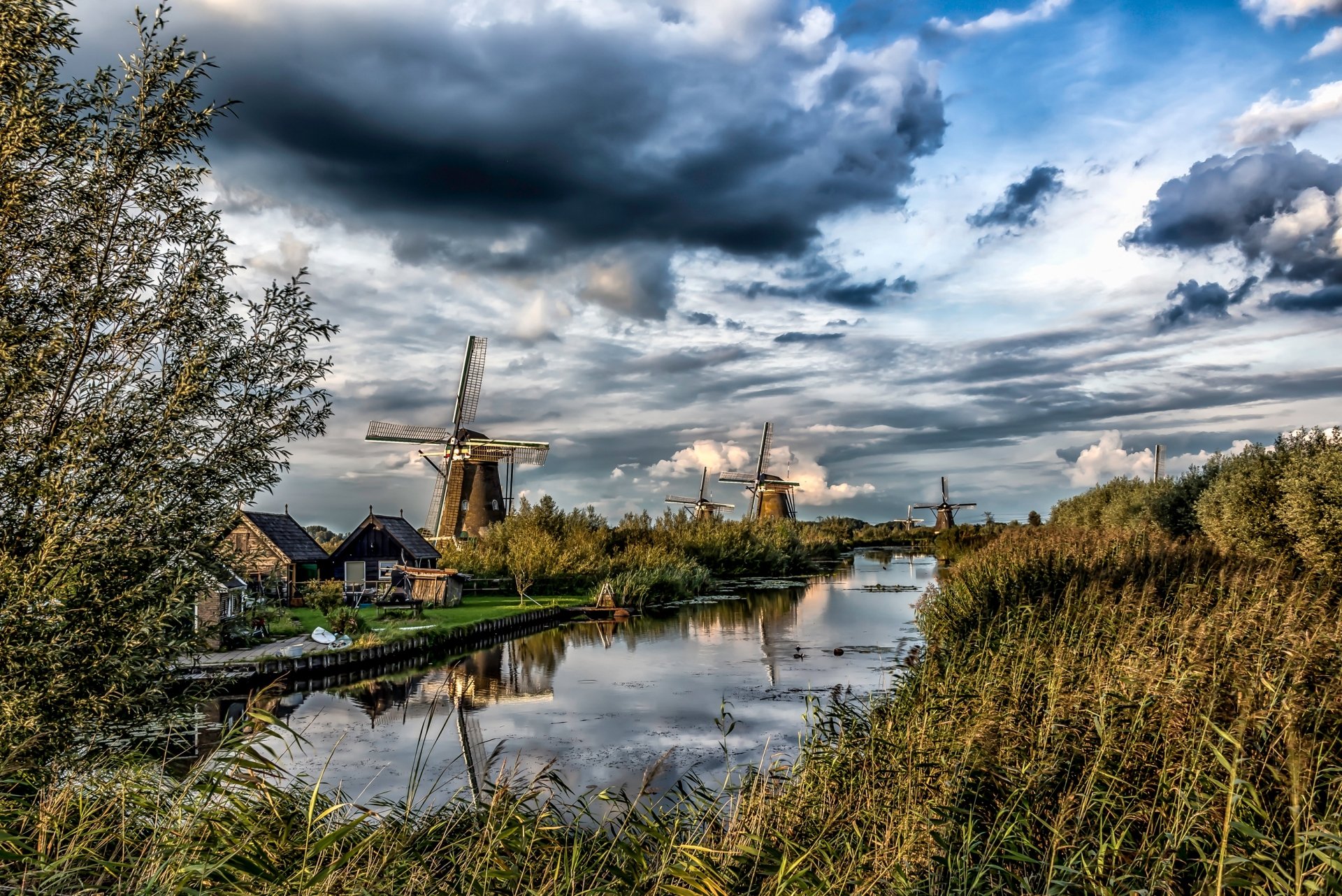 HD desktop wallpaper depicting a serene Dutch river scene with traditional windmills, buildings, and dramatic cloud formations in the Netherlands countryside.
