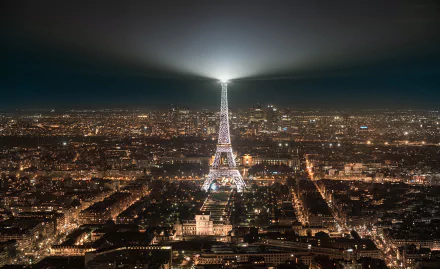Night view of Paris cityscape featuring the illuminated Eiffel Tower monument with its spotlight beam shining across the sky in this HD desktop wallpaper.