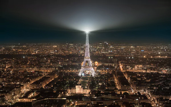 Night view of Paris cityscape featuring the illuminated Eiffel Tower monument with its spotlight beam shining across the sky in this HD desktop wallpaper.