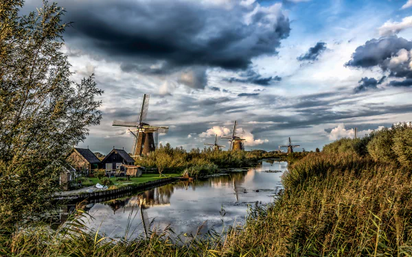 HD desktop wallpaper depicting a serene Dutch river scene with traditional windmills, buildings, and dramatic cloud formations in the Netherlands countryside.