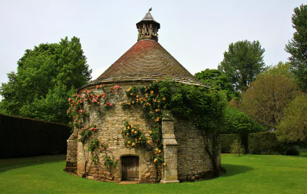 HD desktop wallpaper featuring a stone gazebo adorned with climbing roses in an English park, surrounded by lush greenery and trees.