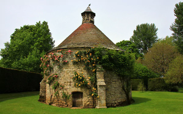 HD desktop wallpaper featuring a stone gazebo adorned with climbing roses in an English park, surrounded by lush greenery and trees.