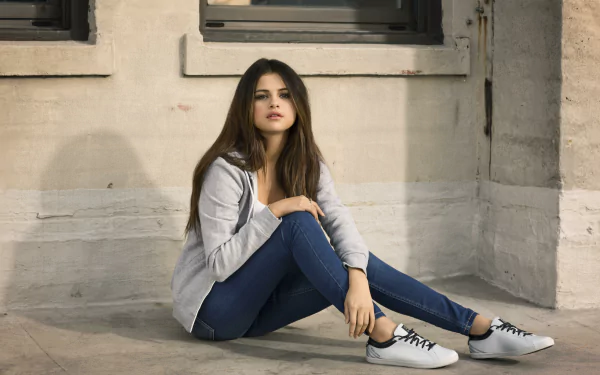 A HD desktop wallpaper featuring Selena Gomez, a brunette singer and actress, sitting against a wall wearing jeans and a casual outfit.