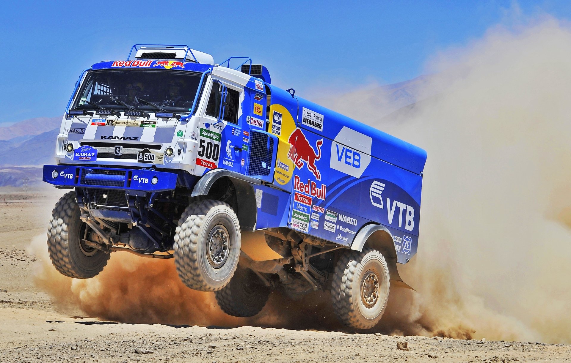 A dynamic HD desktop wallpaper featuring a Red Bull Kamaz rally truck kicking up sand during an intense racing moment. The vehicle's powerful presence is emphasized by its vibrant blue color and various sponsor logos.