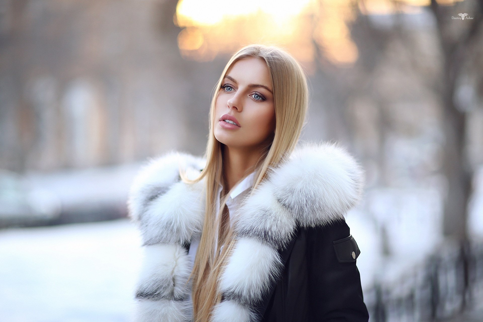 Blonde woman with blue eyes wearing a fur-lined winter coat stands outdoors in soft-focus background, captured in HD with shallow depth of field.