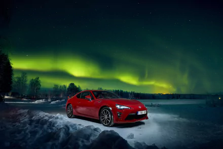 A red Toyota 86 is parked on a snowy road under the vibrant aurora borealis in the night sky. The image is a high-definition desktop wallpaper and background.
