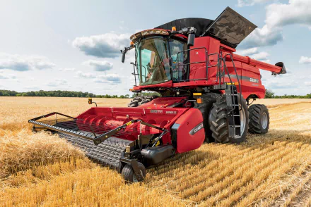 HD desktop wallpaper featuring a red tractor harvesting wheat in a vast summer field under a partly cloudy sky.