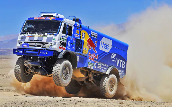 A dynamic HD desktop wallpaper featuring a Red Bull Kamaz rally truck kicking up sand during an intense racing moment. The vehicle's powerful presence is emphasized by its vibrant blue color and various sponsor logos.