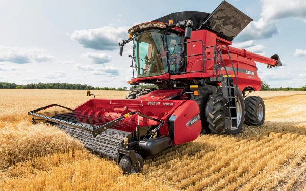 HD desktop wallpaper featuring a red tractor harvesting wheat in a vast summer field under a partly cloudy sky.