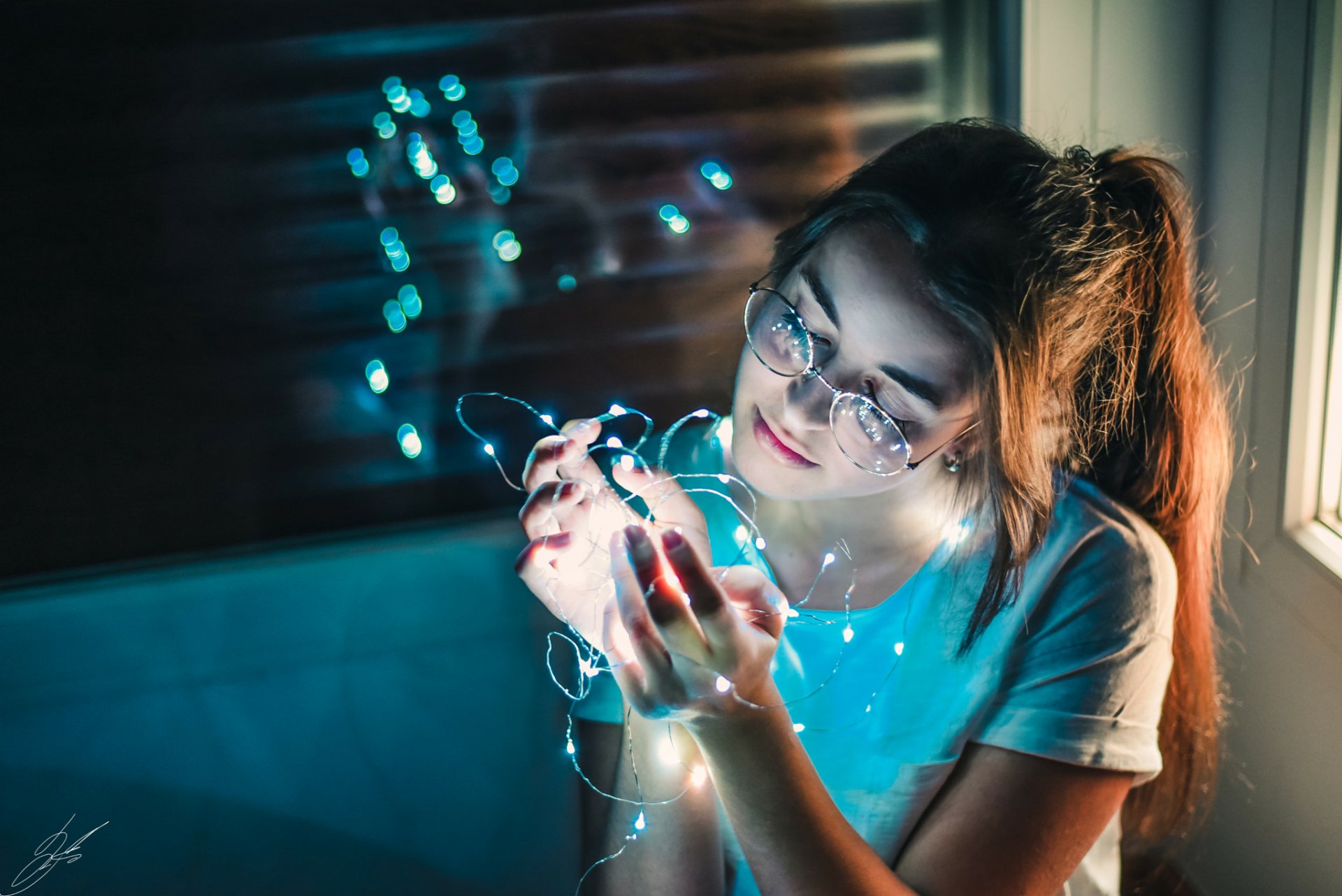 Brunette woman with long hair and glasses gently holding glowing string lights in a softly lit room, captured in a high-definition desktop wallpaper.