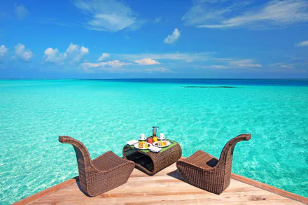 Luxury resort setting with two wicker chairs and a lunch table overlooking a clear turquoise tropical ocean and horizon. HD desktop wallpaper and background image.