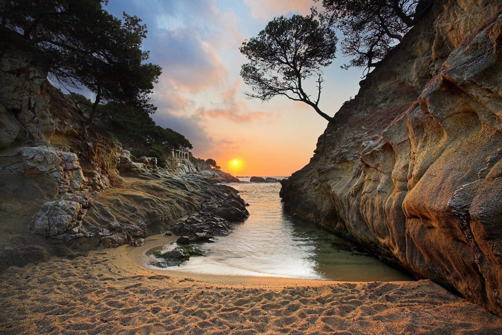 A stunning HD desktop wallpaper of a sunset over a sandy beach in Greece, with a serene coastline, rocky formations, trees, and the sea meeting the horizon.