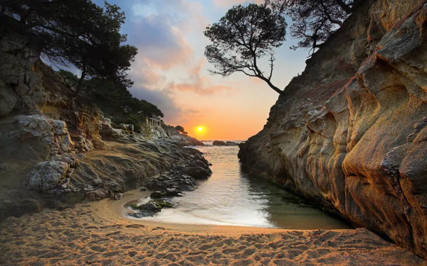 A stunning HD desktop wallpaper of a sunset over a sandy beach in Greece, with a serene coastline, rocky formations, trees, and the sea meeting the horizon.