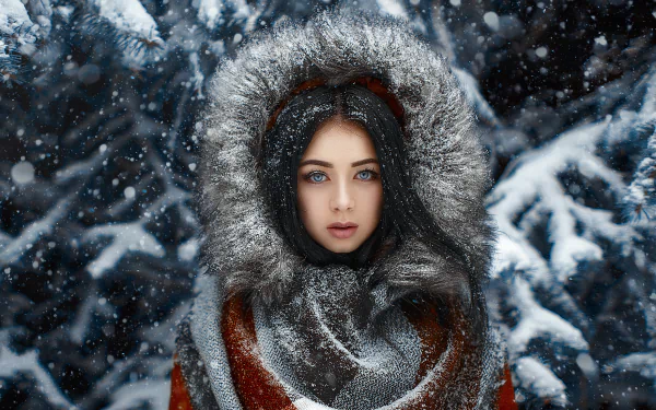 A striking winter HD wallpaper featuring a woman with long black hair and blue eyes, wearing a fur-lined coat amidst falling snow and snow-covered trees.