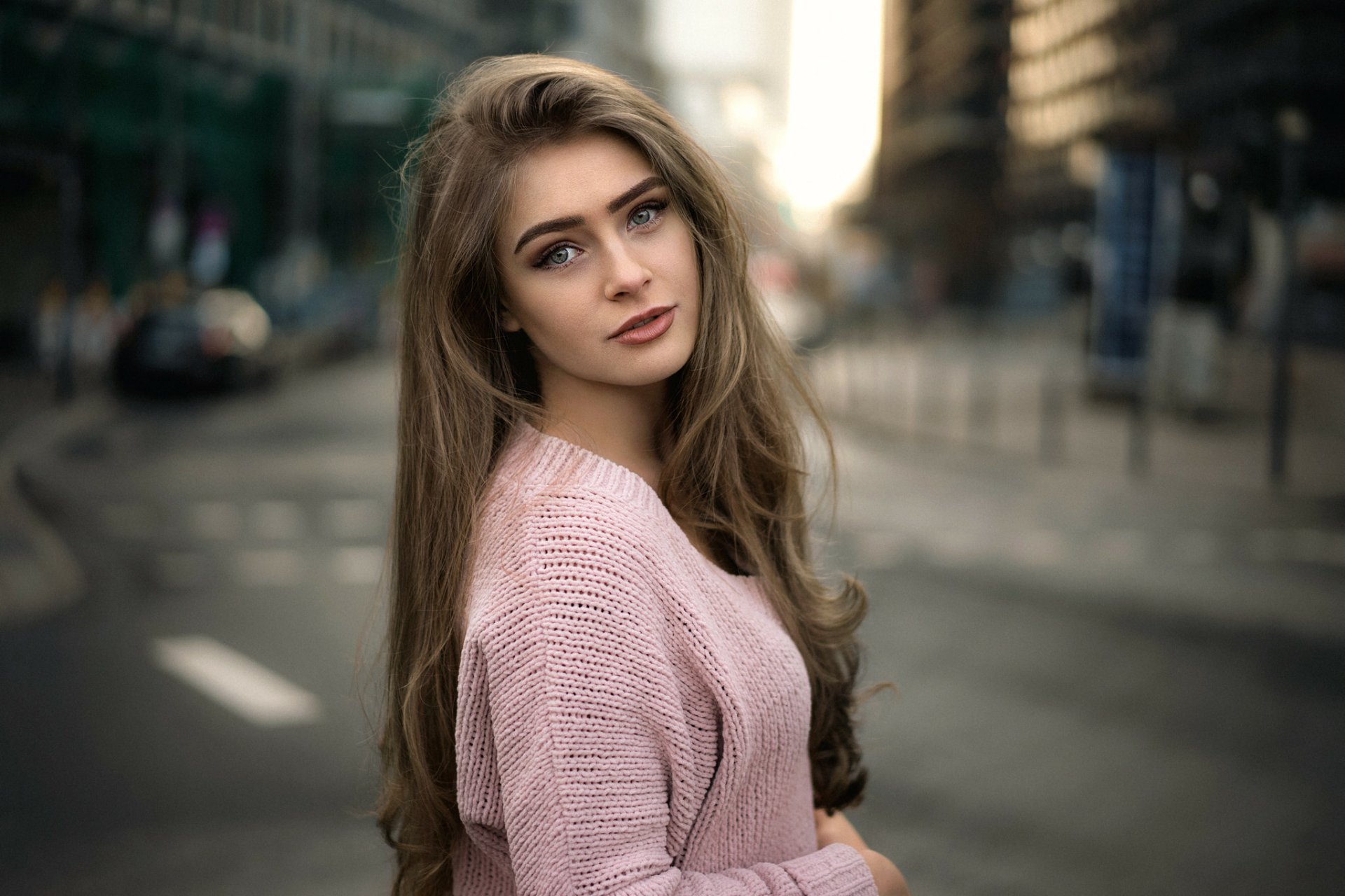 HD desktop wallpaper featuring a brunette woman with long hair, blue eyes, and a pink sweater, captured with a shallow depth of field on a city street background.