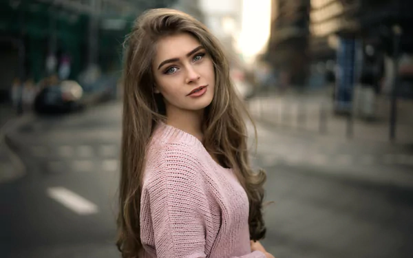 HD desktop wallpaper featuring a brunette woman with long hair, blue eyes, and a pink sweater, captured with a shallow depth of field on a city street background.