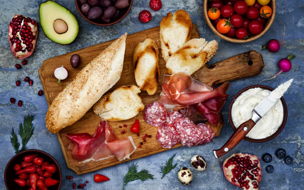 4K Ultra HD PC desktop wallpaper still life of food: sliced salami and cured meat, crusty bread, cheese, avocado fruit, olives and berries artfully arranged on a wooden board.