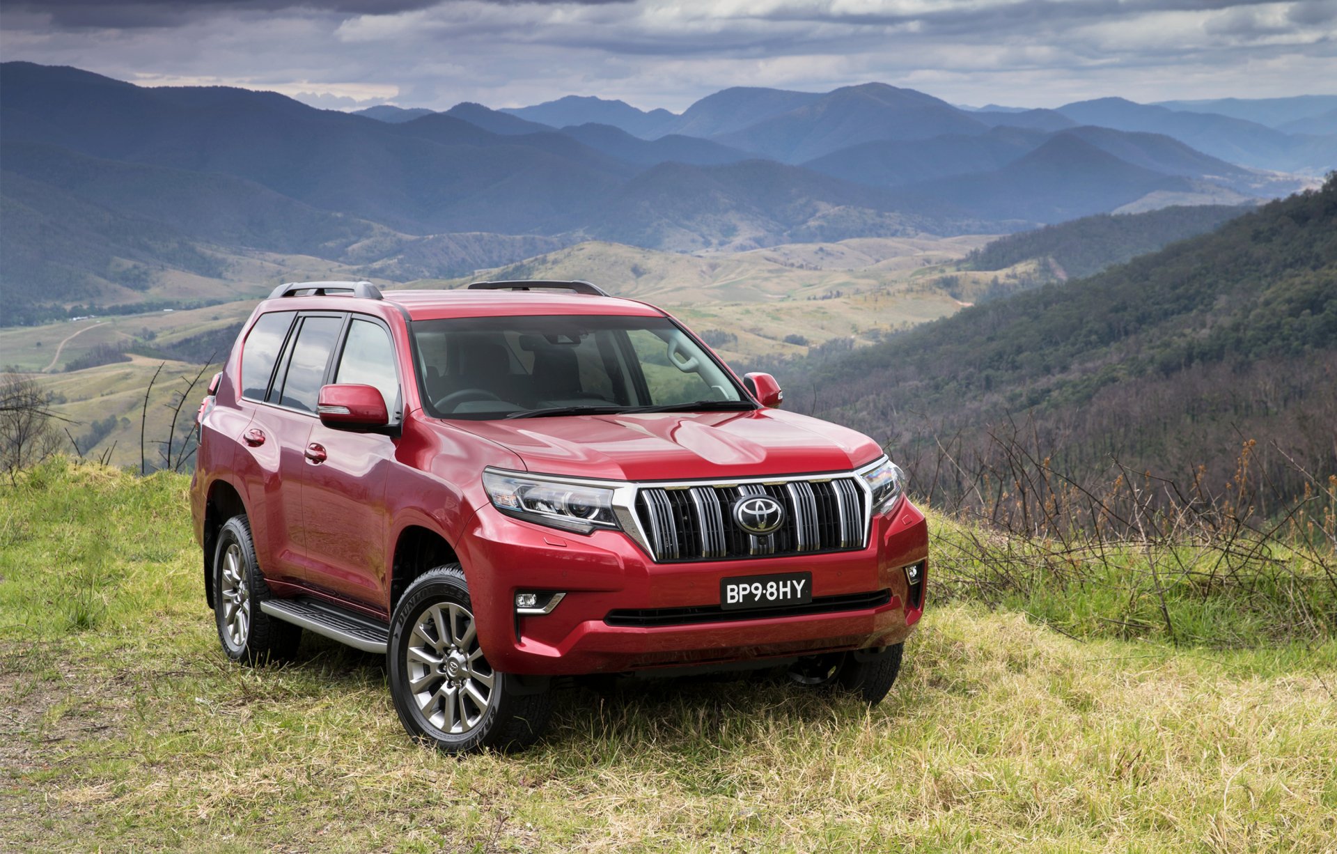 Red Toyota Land Cruiser Prado SUV car parked on a grassy hillside with rolling mountains in the background — 2K Quad HD PC desktop wallpaper.