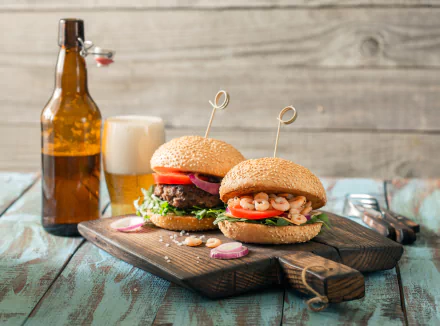 Still life 4K Ultra HD image of two burgers with fresh toppings on a wooden board, accompanied by a frothy beer and an amber bottle on a rustic table.