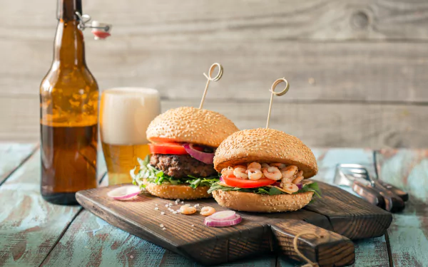 Still life 4K Ultra HD image of two burgers with fresh toppings on a wooden board, accompanied by a frothy beer and an amber bottle on a rustic table.