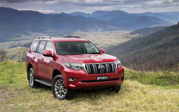 Red Toyota Land Cruiser Prado SUV car parked on a grassy hillside with rolling mountains in the background — 2K Quad HD PC desktop wallpaper.