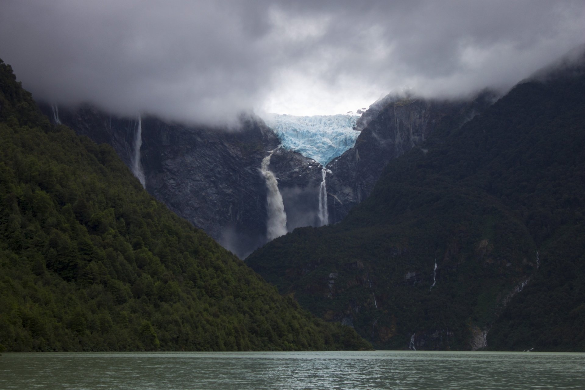 HD desktop wallpaper featuring a serene glacier-fed lake with cascading waterfalls, surrounded by lush green mountains under a cloudy sky.