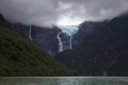 HD desktop wallpaper featuring a serene glacier-fed lake with cascading waterfalls, surrounded by lush green mountains under a cloudy sky.