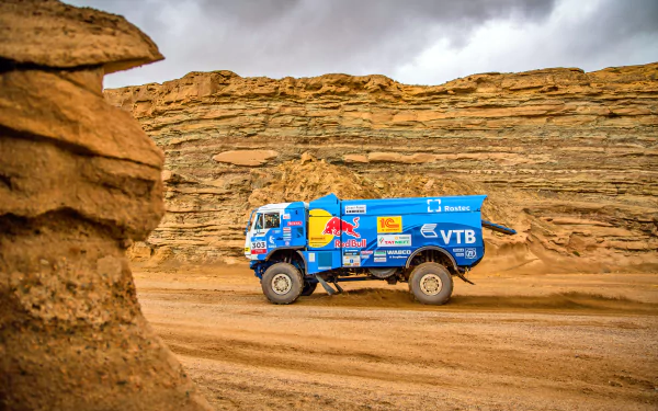 A blue rally truck speeds through a rugged desert landscape with layered rock formations under a cloudy sky, captured in 4K Ultra HD for a dynamic PC desktop wallpaper.