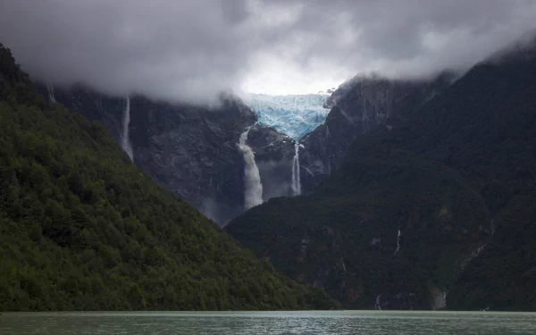 HD desktop wallpaper featuring a serene glacier-fed lake with cascading waterfalls, surrounded by lush green mountains under a cloudy sky.