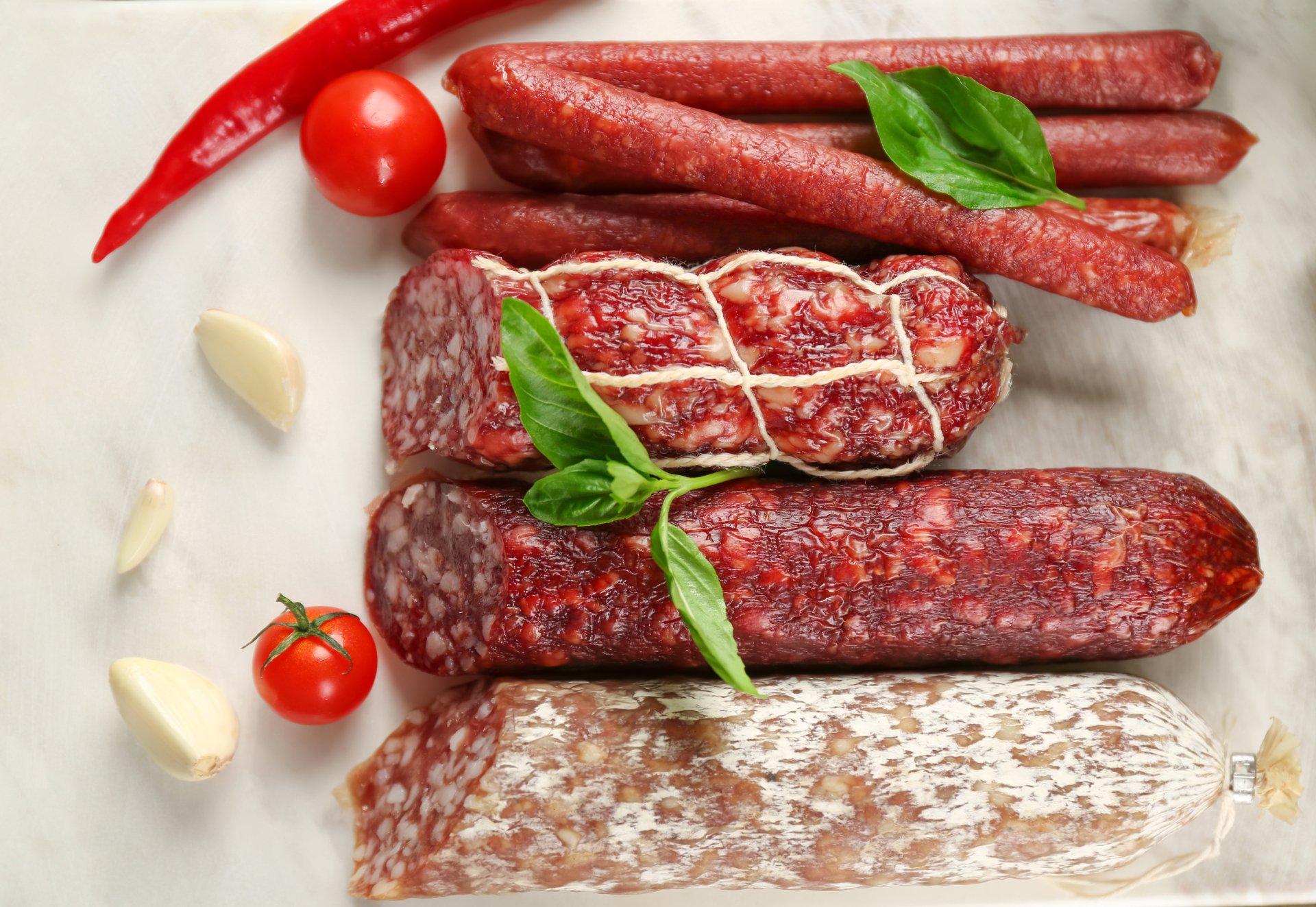 4K Ultra HD still life of assorted sausages and salami with fresh tomatoes, garlic cloves, and basil leaves on a light surface, showcasing rich colors and textures.