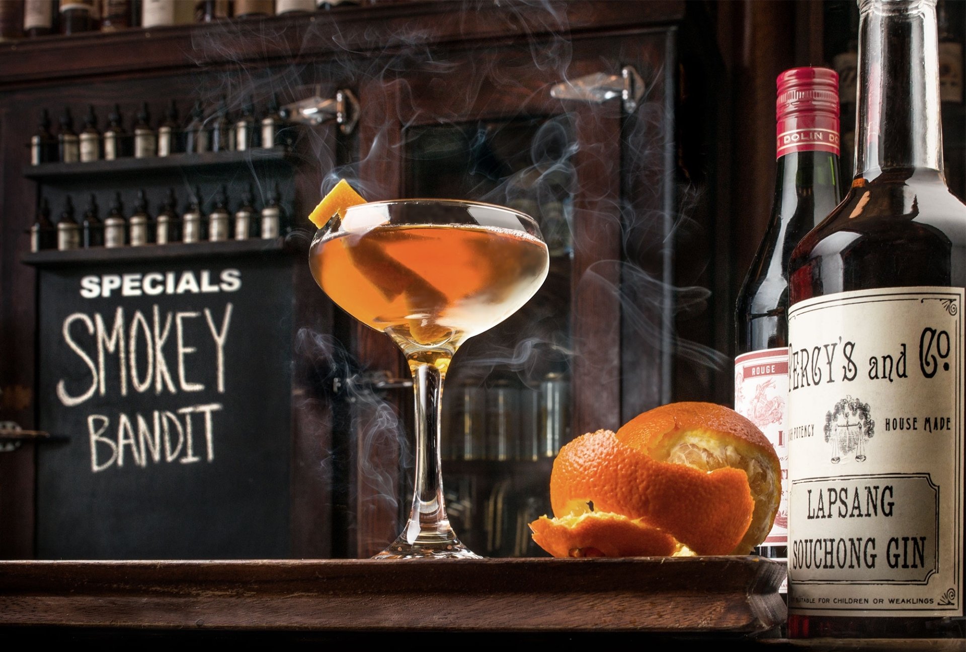 HD desktop wallpaper featuring a smoky cocktail in a glass garnished with citrus peel, with oranges and a bottle of Lapsang Souchong Gin on a bar counter, labeled Smokey Bandit.