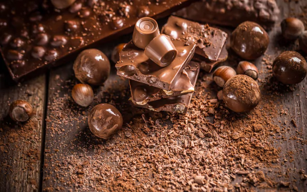 HD desktop wallpaper featuring assorted chocolate sweets and whole hazelnuts on a rustic wooden surface, with chocolate shavings scattered around.