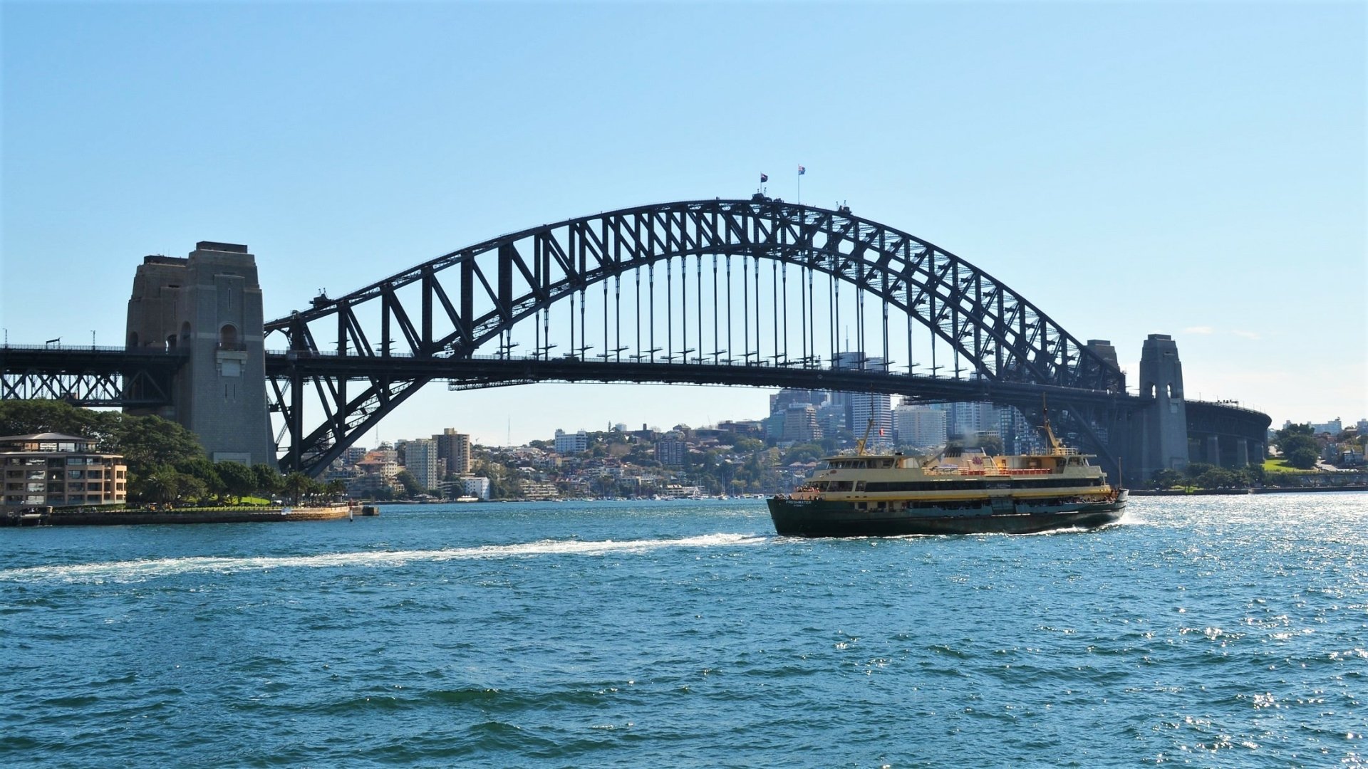 HD desktop wallpaper of a ferry crossing Sydney Harbour with the iconic Sydney Harbour Bridge in the background on a clear, sunny day.