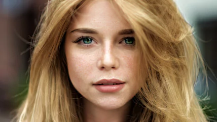 Close-up HD portrait of a blonde woman with blue eyes, freckles, and soft waves, showcasing a natural and striking model face against a blurred background.